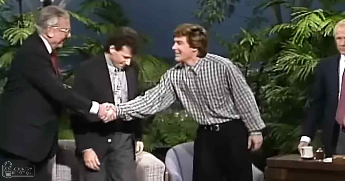 Two adult males shaking hands in a well-lit indoor setting with greenery, possibly for a stage or studio.