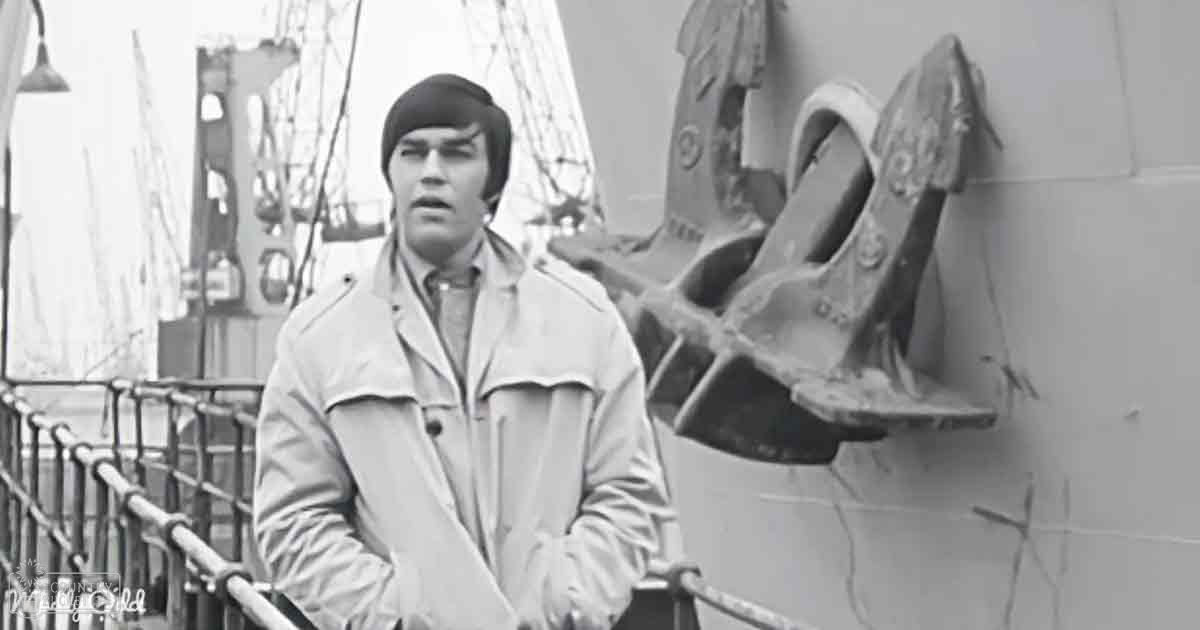 A young adult male wearing a beanie hat, trench coat, and collared shirt stands outdoors on or near a ship in an urban harbor setting with rigging visible in the background.