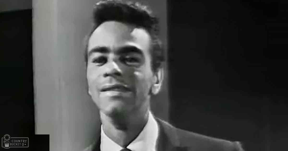 A young adult male in a dark suit jacket and white shirt stands indoors against a dark curtain backdrop, possibly during daytime with artificial light.