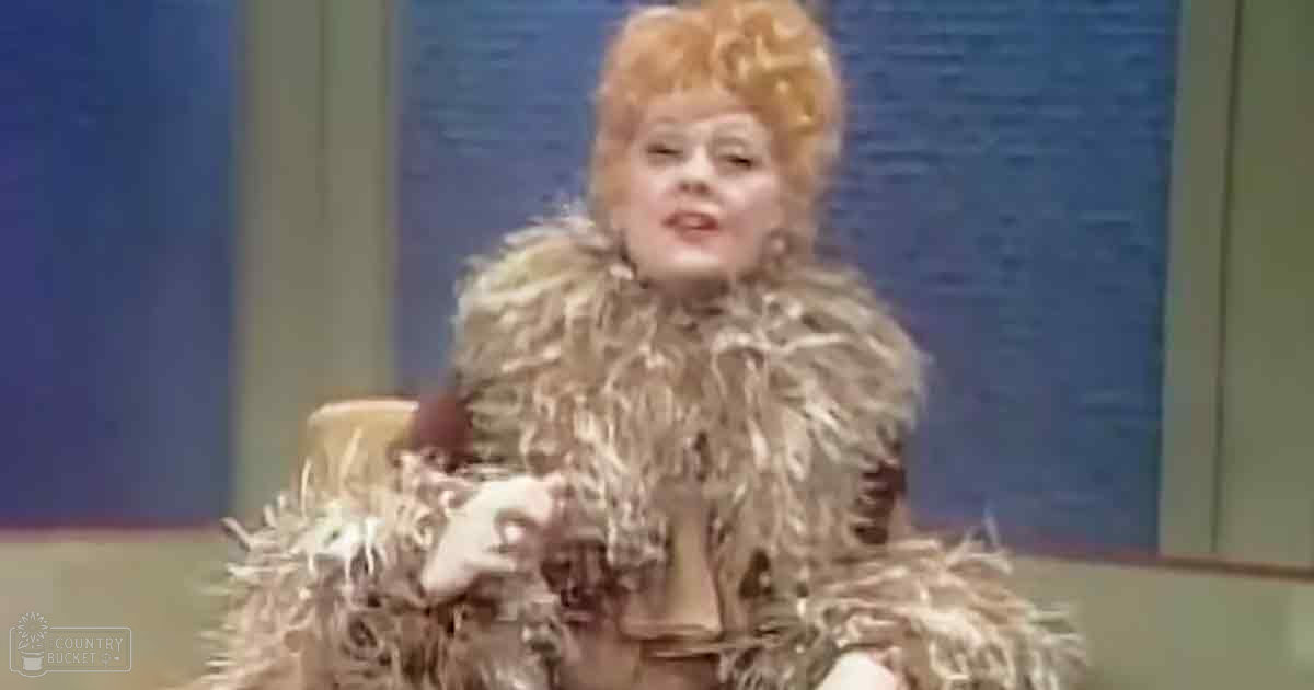 A middle-aged female wearing a luxurious light brown fur coat with ruffled details sits on an elegant chair in front of a blue curtain, likely in a studio or stage setting.
