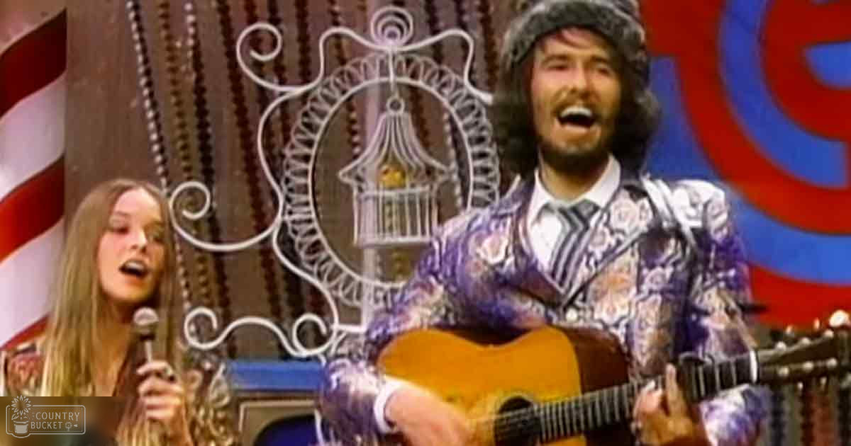 A musical performance scene with two individuals on stage; one young adult female in a patterned purple and white top, middle-aged male in a loose-fitting paisley shirt, decorative birdcage with a yellow bird behind the male subject, American flag in the background, suggesting an indoor venue from the 1960s or 1970s.