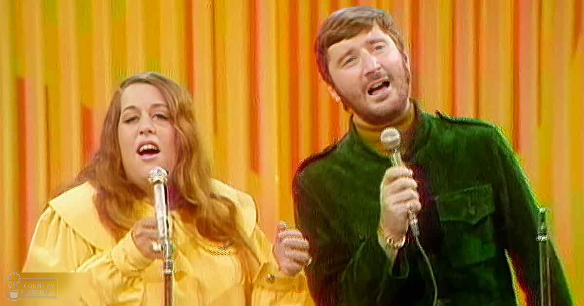 A musical performance scene with a young adult female wearing a shiny, bright yellow dress holding a microphone and an adult male singer in a green velvet jacket also holding a microphone against a backdrop of orange and yellow striped curtains.