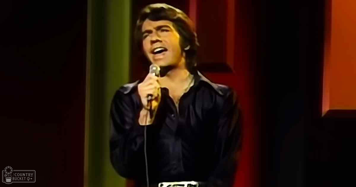 An adult male singer on stage, wearing a dark-colored long-sleeved shirt with an open collar and holding a microphone in front of curtains and stage lighting.