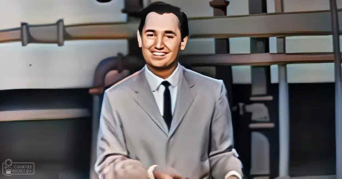 Adult male in light gray suit jacket with notched lapels, standing indoors near a staircase with wooden railings. Bright daytime lighting illuminates the scene.