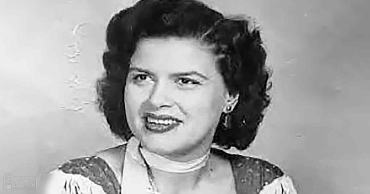 A middle-aged woman in a patterned garment with intricate designs and a high neckline stands indoors against a plain light-colored background, suggesting daytime or early evening.
