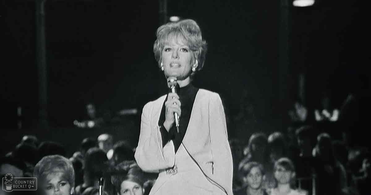 An adult female performer in a white blazer and black shirt, holding a microphone, stands on stage with audience seating behind her.