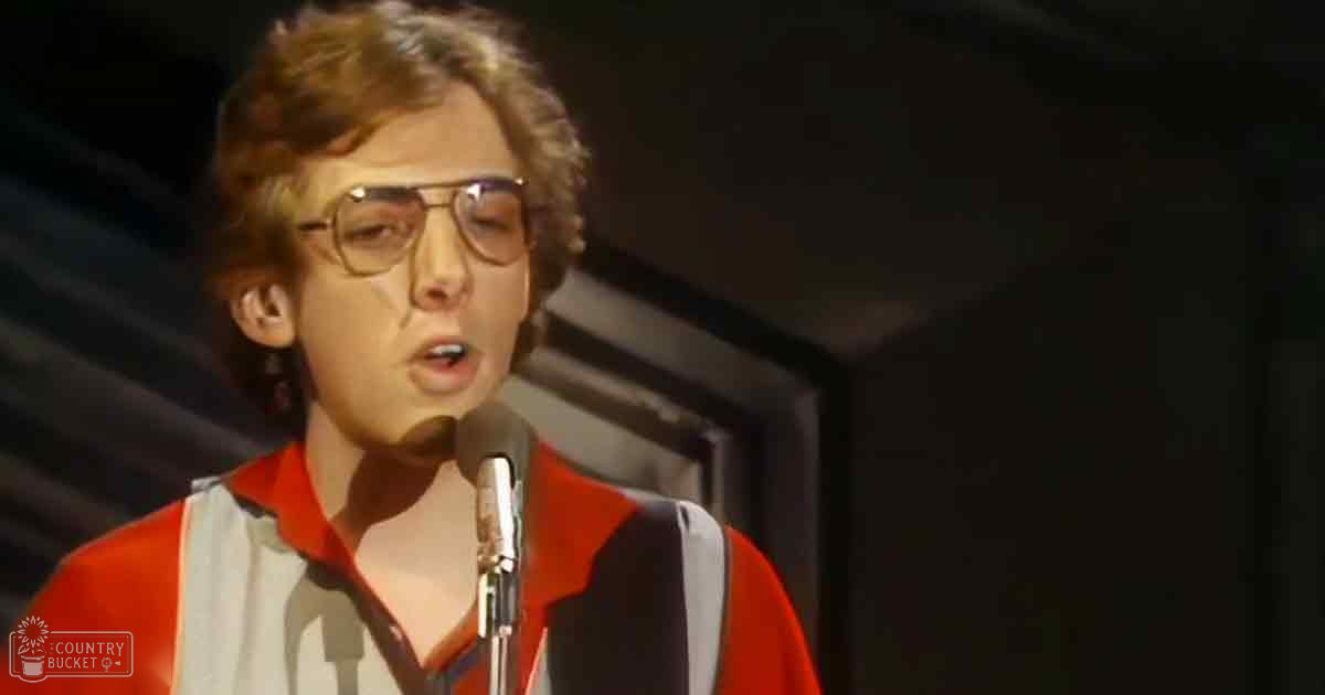 A young adult male wearing a red and white striped, collared, long-sleeved shirt with thick-framed glasses sings into a microphone on stage in dimly lit indoor setting.