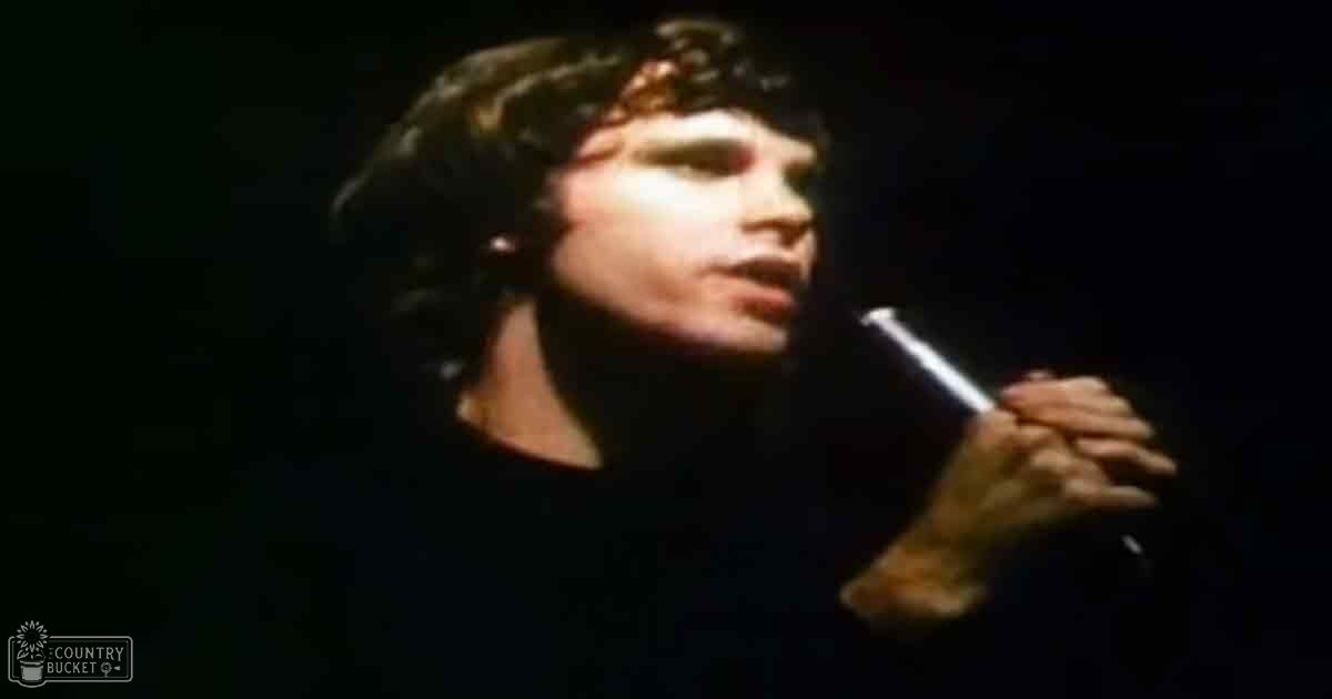 A young adult male singer performing on stage, wearing a dark-colored t-shirt or long-sleeved shirt with a crew neckline and short sleeves, holding a microphone close to his mouth. The setting appears to be an indoor venue with dim lighting focused on the performer during nighttime.
