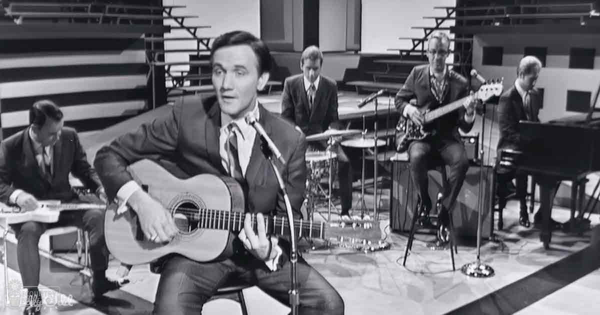 Studio setting with musicians playing instruments, including a man with an acoustic guitar seated on a stool.