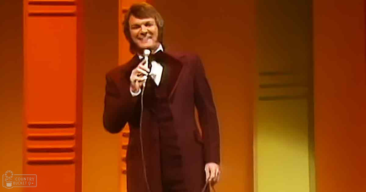 An adult male performer wearing a tailored long black coat and white shirt with a bow tie, holding a microphone, likely in a mid-20th century theatrical setting with orange and yellow stage lighting.