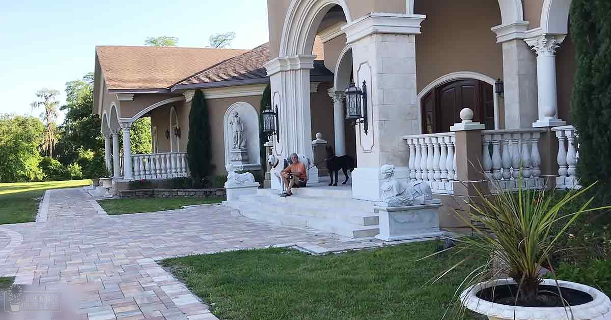 A medium-sized dog with a black coat sits casually in front of a white-porched entrance surrounded by lush greenery and paved walkways, suggesting spring or summer.