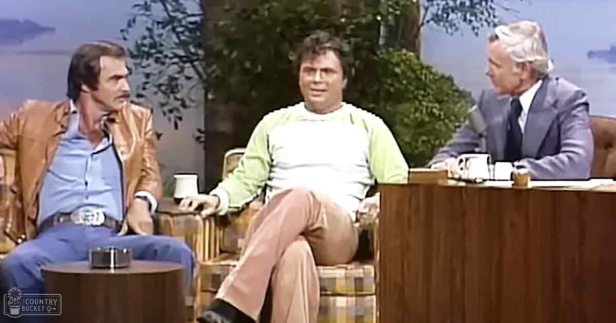 A talk show set outdoors with three adult men, one older and two younger, seated on armchairs under natural light among trees.