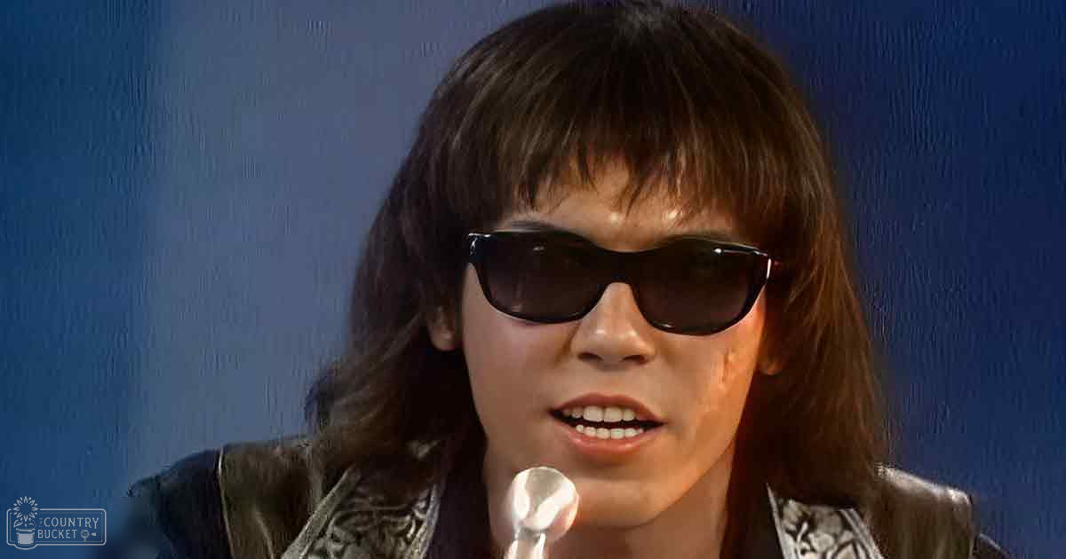 Young adult male in a relaxed-fit, collarless leather jacket with intricate floral or paisley patterns and sunglasses stands indoors near a microphone against a blue wall.