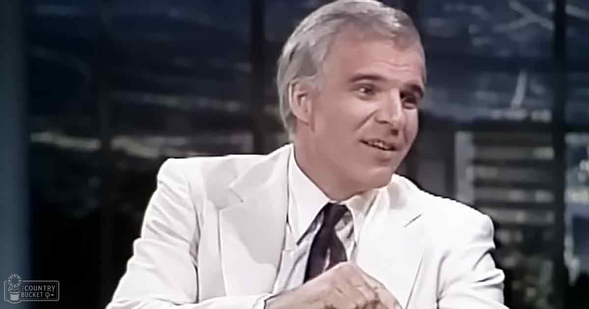 Middle-aged to older adult male in a professional studio setting, wearing a white suit jacket with a notched lapel and dark patterned tie.