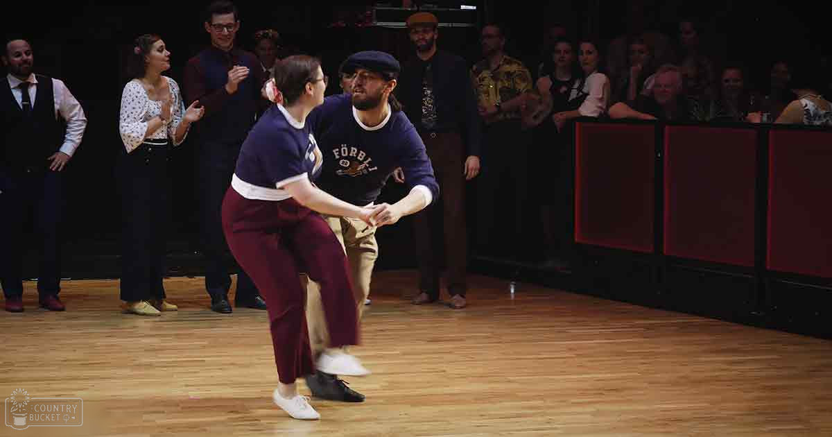 A dance duo performing in a studio or performance space with spectators watching.