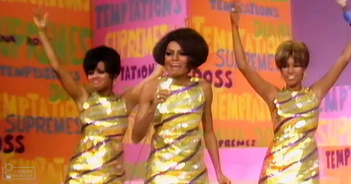 Three young adult females wearing matching sleeveless gold dresses with vertical stripes perform on a brightly lit stage with 'Supremes' text in the background.