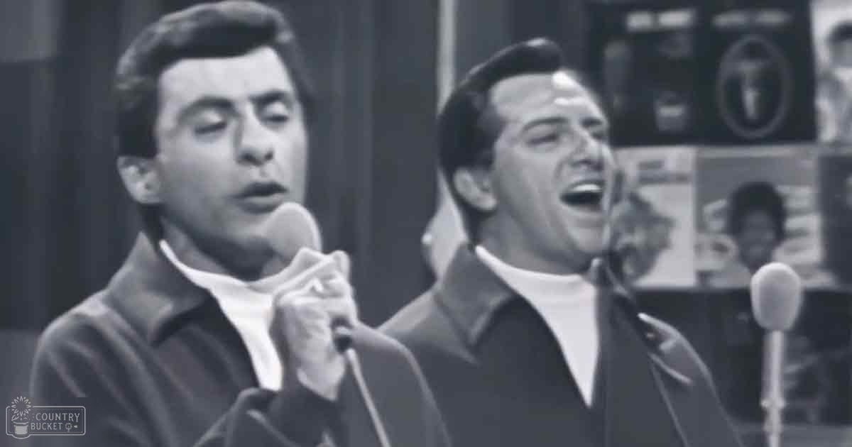 Two young adult males singing in a recording studio setting with microphones and control panel visible, wearing dark jackets and suits.