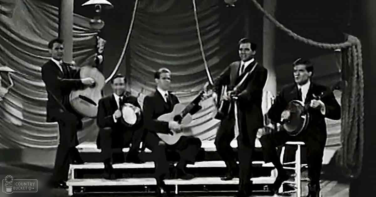 A mid-20th century musical performance with musicians in dark suits playing string instruments on a stage with draped fabric and even lighting.