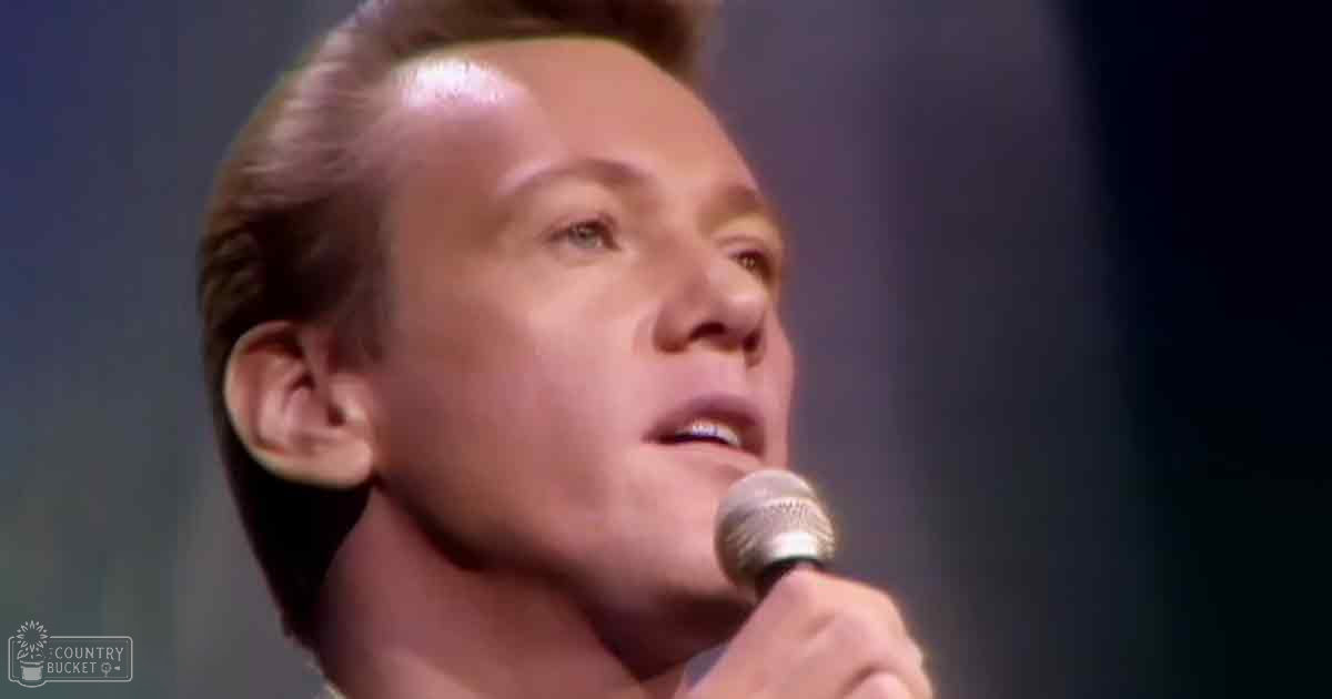 A middle-aged man in a dark, semi-formal shirt sings into a microphone with 'Madly Odd' text faintly visible. The setting is dimly lit on stage.
