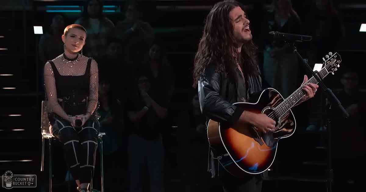 A young adult female performer wearing a black lace dress and choker necklace plays guitar while an adult male singer holds his acoustic guitar and sings into a microphone, set against a dark studio background with blurred audience figures in the distance.