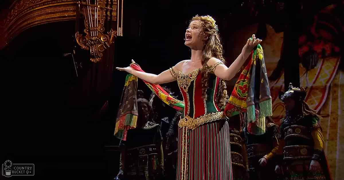 A female performer in an ornate, historical costume with red, green, black, and gold colors on a grand stage resembling a Renaissance or Baroque palace.