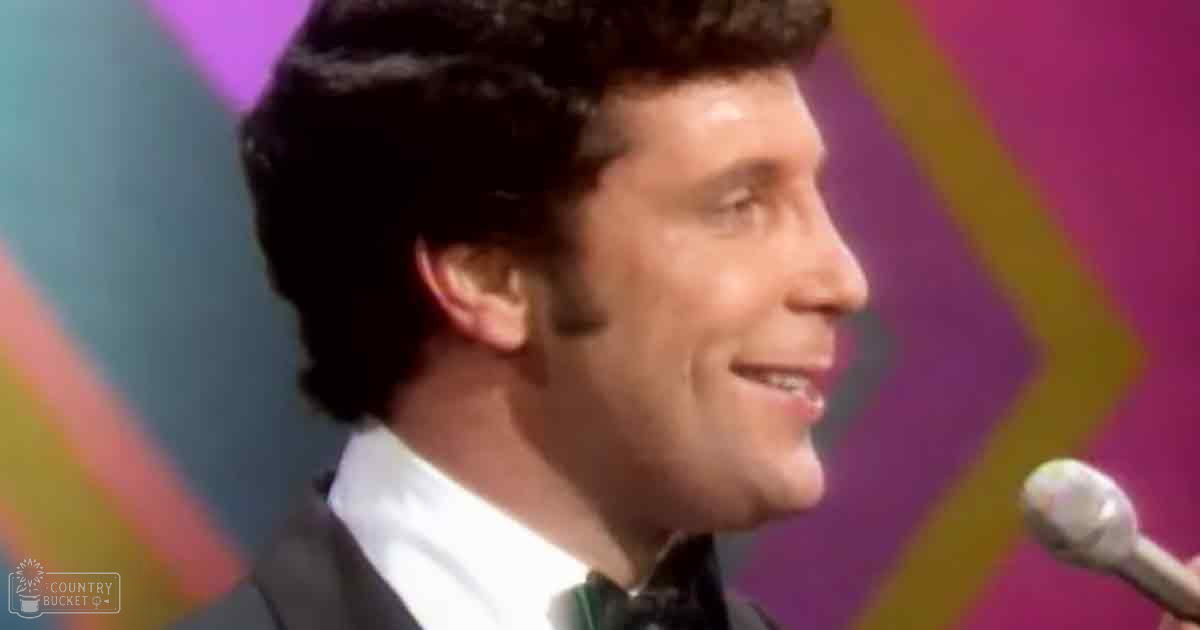 A young adult male dressed in a 1970s-style dark suit jacket, white dress shirt, and black bow tie stands on stage near a microphone with 'Madly Odd' text visible. The setting is vibrant and abstract indoors under artificial lighting.
