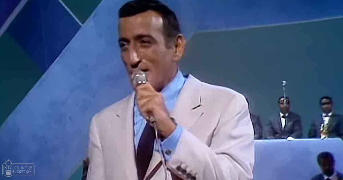 A male performer singing into a microphone on stage; he wears a light-colored suit jacket and white shirt with a dark tie. In the background are two adult males, one seated in a dark suit playing an instrument.