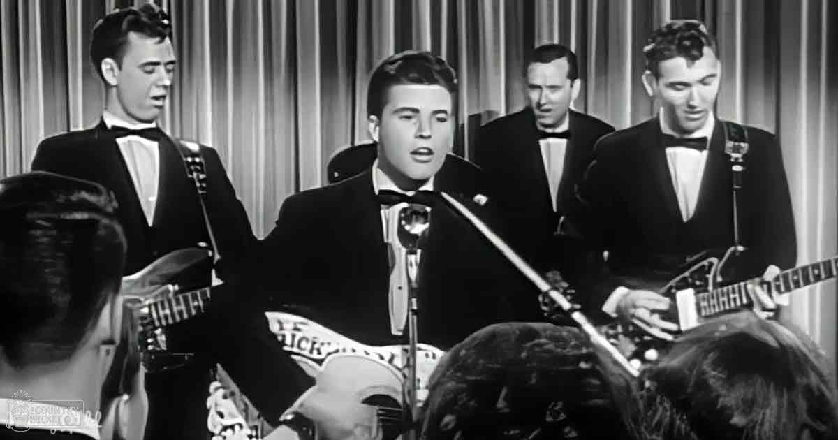 A young adult male singer wearing a dark suit jacket and white shirt with a bow tie performs on stage in front of a band, with vertical striped curtains as the background.