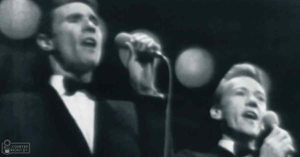 Two male performers in dark suits singing into microphones on an indoor stage with mid-20th century ambiance.