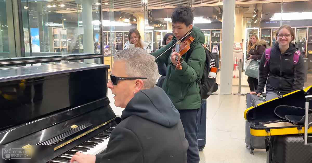 A middle-aged to older adult man plays a black upright piano with a gold plaque in an airport setting, surrounded by young adults and luggage.