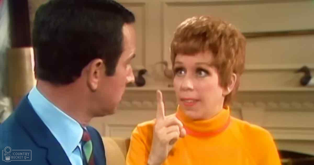 An adult female wearing a bright orange turtleneck sweater stands indoors, surrounded by wooden cabinetry in what appears to be a domestic kitchen setting during daytime.
