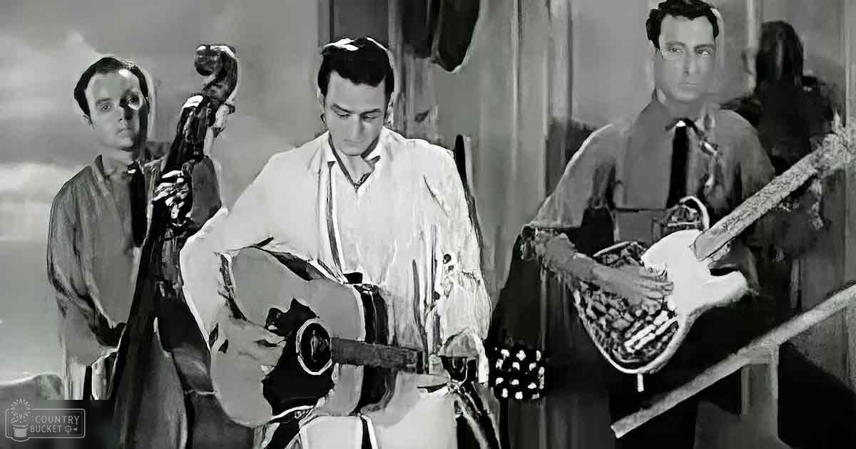A rustic indoor setting with three adult males playing guitars; one in a light shirt and another in a frayed white button-up shirt, both standing on a wooden structure holding their instruments.