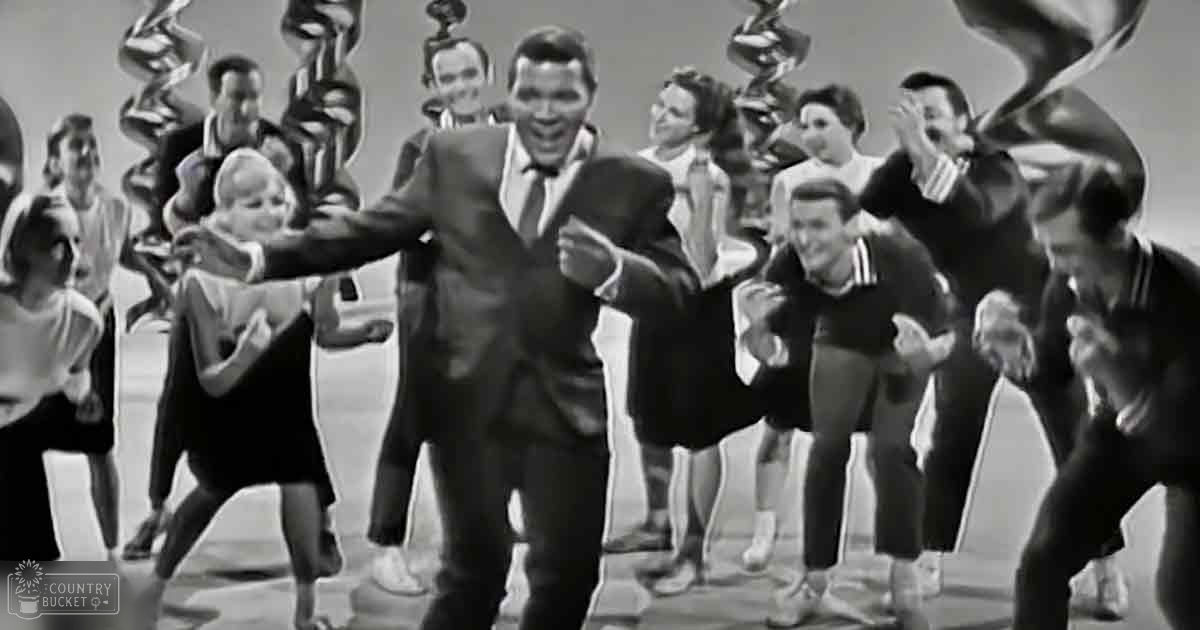 A man in his adult years, dressed formally in a dark suit with a white shirt and bow tie, stands amidst children wearing casual clothes at an indoor event or performance setting with abstract sculptures.