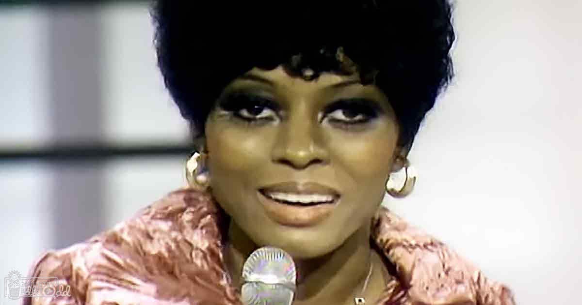An adult female performer wearing a textured pink and brown garment, holding a microphone near her mouth against an indoor stage backdrop.