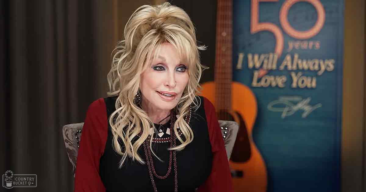 A middle-aged adult female wearing a black, intricately patterned top with beadwork and long-sleeved red pants indoors. A chair with ornate detailing and a guitar leaning against the wall are visible in the background.