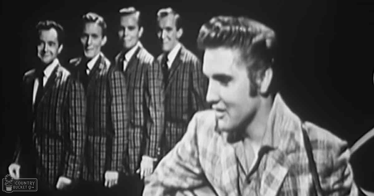 A young adult male, likely in his late teens or early twenties, plays an instrument (possibly a guitar) against a backdrop of adults dressed in mid-20th century plaid suits and jackets, under dimly lit studio conditions with black-and-white photography.