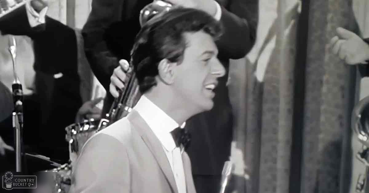 A young adult male singer wearing a white suit jacket and bow tie performs on stage with other individuals in formal attire.