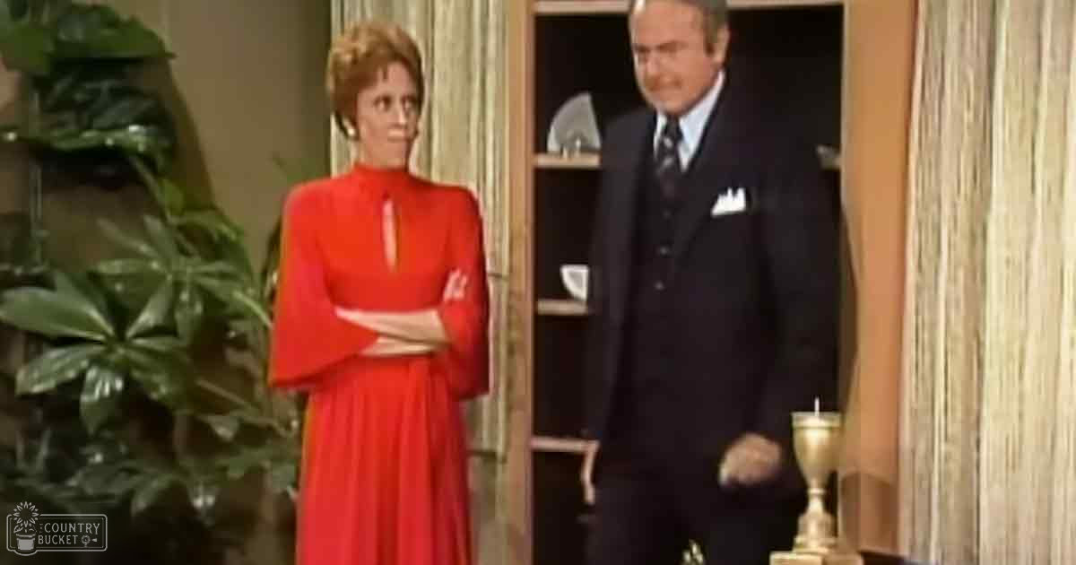 A woman wearing an orange bell-sleeve dress stands in a living room with plants and furniture, while a man in a dark suit is partially visible behind her.