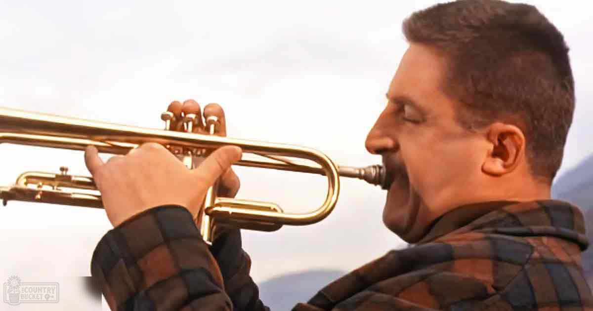 An adult male playing a trumpet outdoors under cloudy skies with mountains in the background.