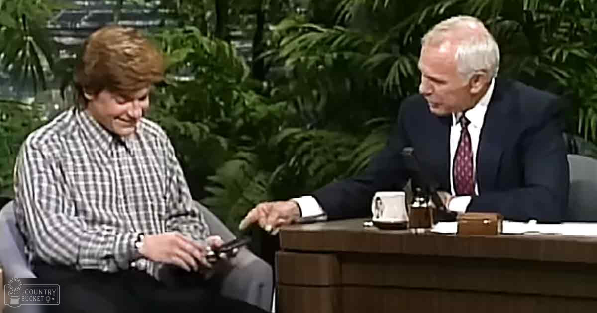 A young adult male in a plaid shirt interview setting with an older adult male in a formal suit jacket on stage with indoor plants and natural elements.
