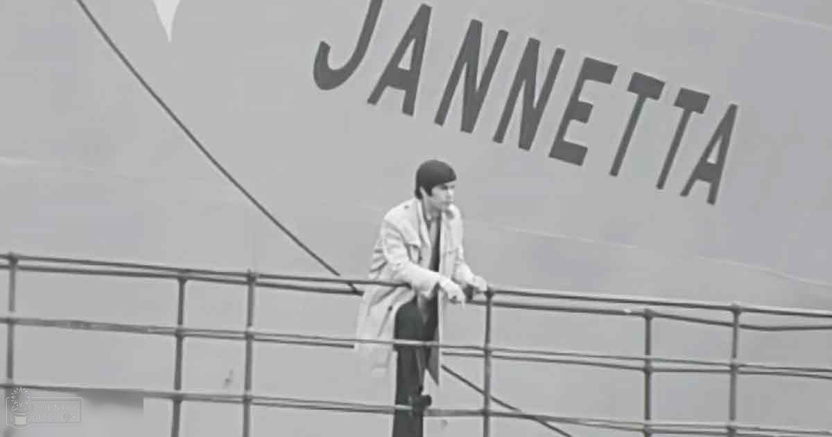 A young adult male stands on the deck of a ship named 'Jannetta' at sea with clear skies in black and white.