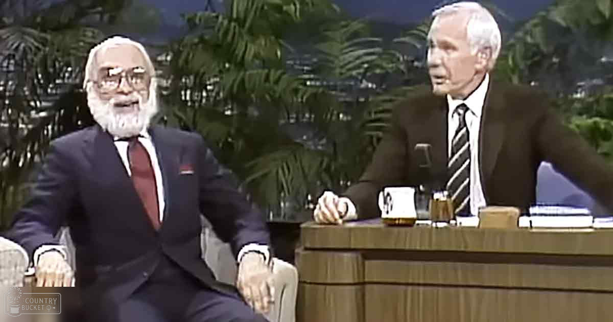 A talk show host and guest in a television studio set. The host wears an older adult male, dark suit jacket with white shirt and red tie, glasses, while the guest is middle-aged to older adult, dressed formally in a striped tie and dark trousers.
