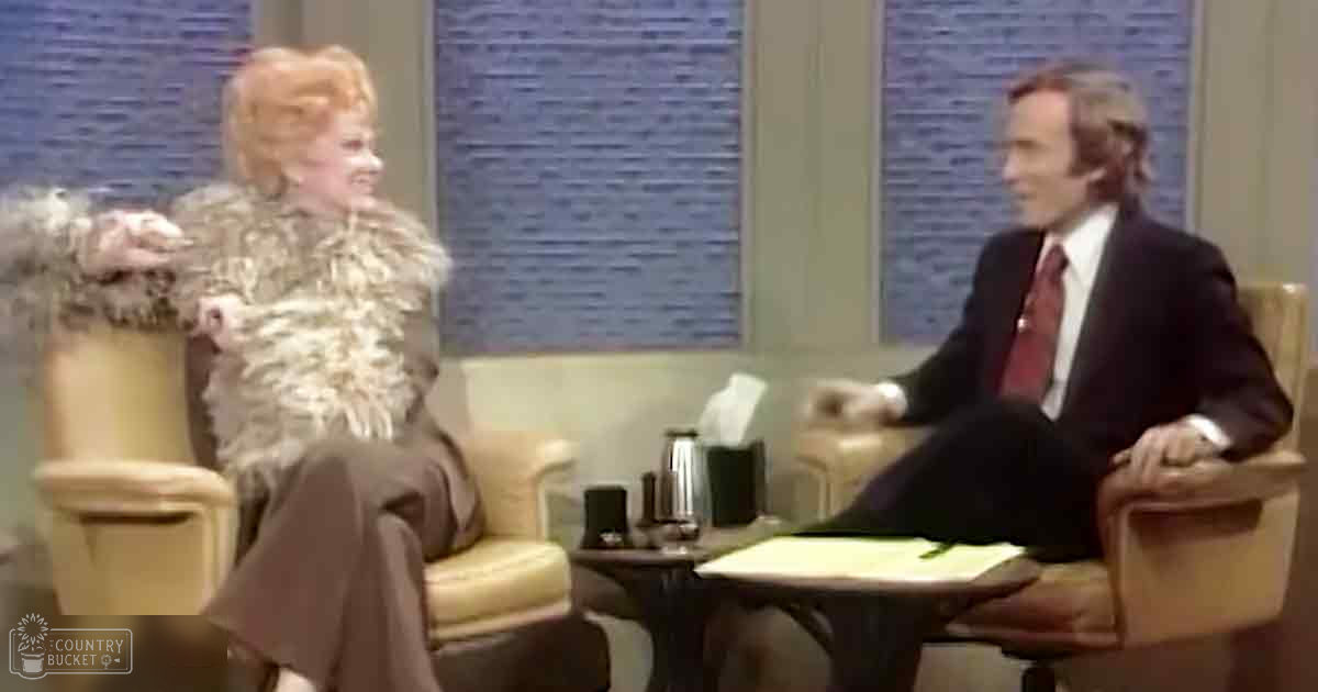 Two talk show hosts in mid-20th century style attire seated on beige armchairs with dried flower decorations, simple studio background