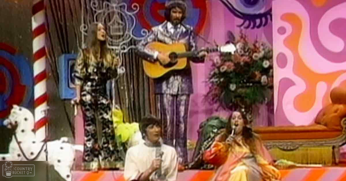 A vibrant, psychedelic musical performance in an indoor setting with swirling patterns and a decorative wrought iron gate; a standing woman in a black and white patterned outfit holds a microphone, alongside a seated man wearing a white shirt and dark trousers holding another microphone, and a seated woman in orange and yellow patterned pants with a scarf or shawl draped over her shoulders also holding a microphone.
