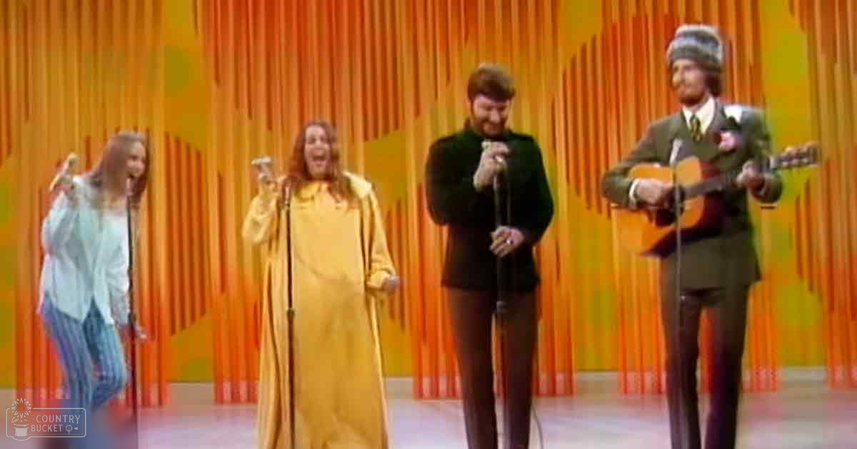 A mid-20th-century indoor musical performance with four adult performers on stage, each holding a microphone or instrument, against an orange and yellow striped backdrop with bright lighting.