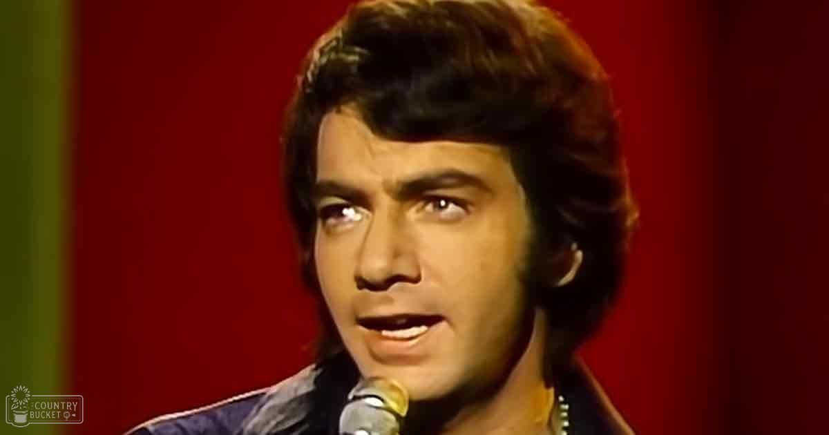 A young adult male wearing a dark-colored, collared, fitted jacket stands on stage with a microphone in front of him. The background is red, suggesting an indoor performance environment.