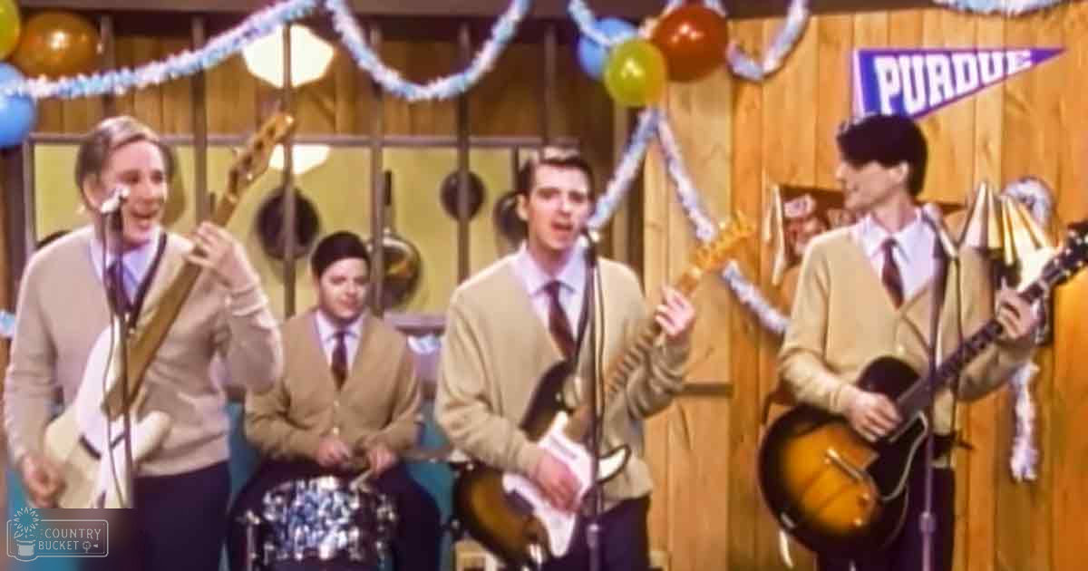 Young adult males performing musical instruments on stage at Purdue University during a festive event with wooden-paneled walls, balloons, and streamers.