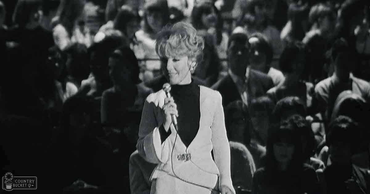 An older adult female performer in a white blazer at an indoor concert venue with seated audience members in casual attire.