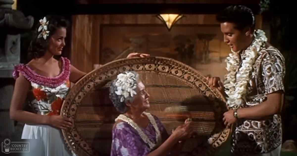 A tropical film set with three individuals wearing distinct Hawaiian attire, holding a woven basket in front of wooden furniture, under natural daylight.