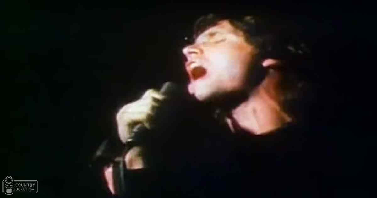 A young adult male singer wearing a dark, collarless cotton shirt performs on stage under dim lighting at night.